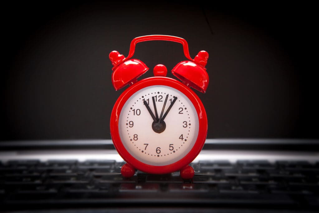 A red retro alarm clock sits on a computer keyboard. The clock shows the time as 12:00, with the hour and minute hands pointing straight up. The background is dark, emphasizing the bright red color of the alarm clock.