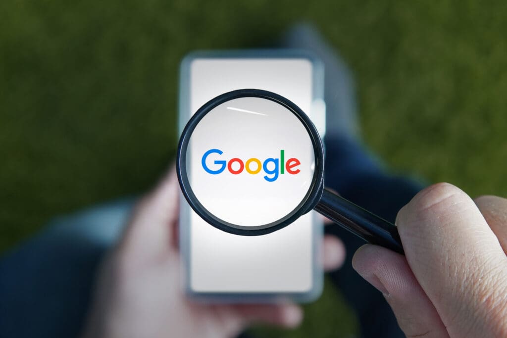 A person holds a magnifying glass over a smartphone displaying the Google logo. The background is blurred, and the logo appears magnified through the glass.