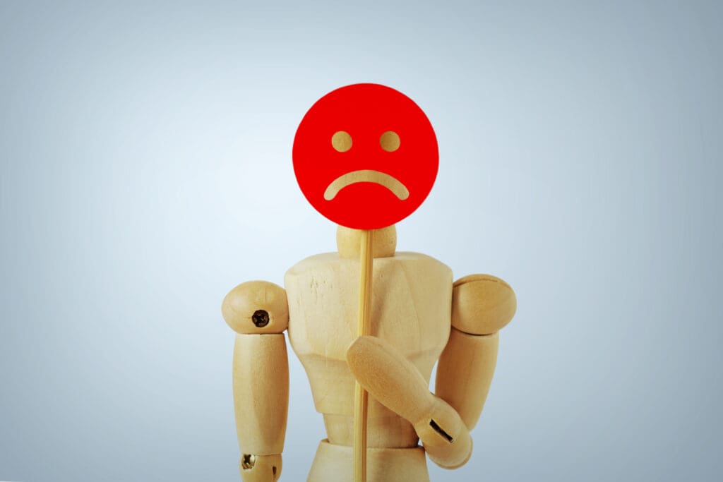 A wooden mannequin holds a stick with a red circle featuring a white sad face, symbolizing unhappiness. The background is pale blue, giving the scene a simple and neutral look.