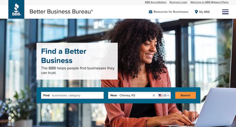 A woman with curly hair smiles as she works on a laptop at a desk. Behind her, sunlight streams through a large window. Text on the image reads, "Find a Better Business," with tips on how to remove a BBB complaint for businesses near Cheney, KS. A search bar is displayed below.