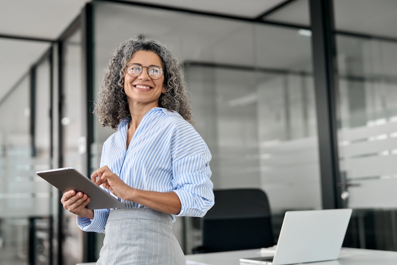 A woman with curly gray hair and glasses, wearing a blue striped shirt, smiles while holding a tablet. She stands in a bright office with glass walls and a laptop on a desk nearby.