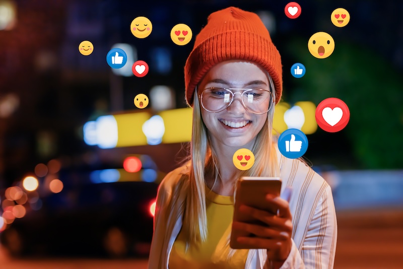 A girl wearing a hat looking at her phone with positive social icons floating around her.