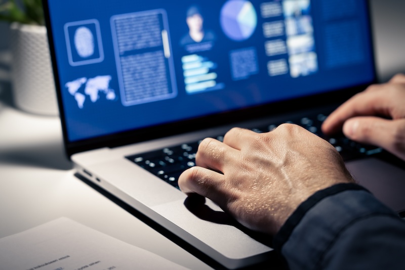 A person typing on a laptop, displaying a screen with text, charts, and a fingerprint icon. The dim lighting suggests a focused, possibly cybersecurity-related task to delete my information online.