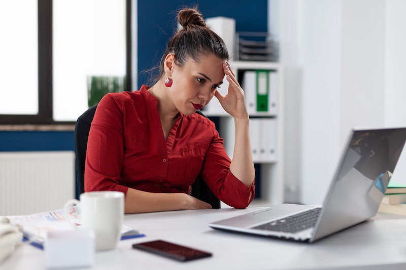 A woman in a red shirt sits at a desk, looking frustrated as she grapples with online mistakes on her laptop. One hand holds her head. Nearby, a smartphone and coffee mug rest on the desk. The background features a window and office shelves.