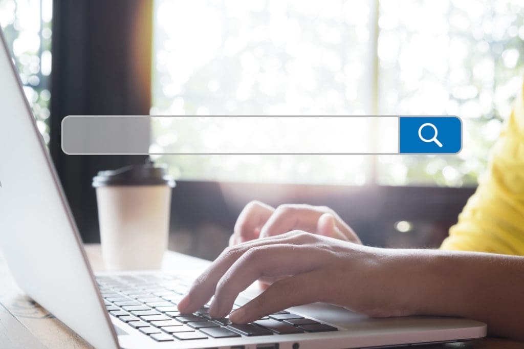 Hands typing on a laptop keyboard with a predictive search bar overlay on the image. A takeaway coffee cup is nearby, and sunlight filters through a window in the background.