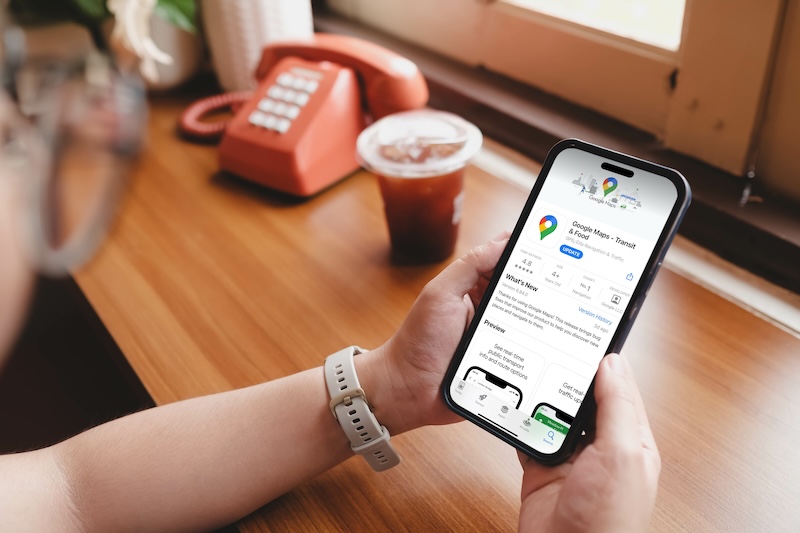 A person holds a smartphone displaying the Google Maps app page in an app store, highlighting its seamless integration with Yelp business reviews. The background features a wooden table with a red rotary phone and a plastic cup filled with iced coffee near a sunlit window.