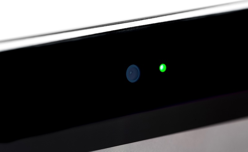 Close-up of a laptop's webcam with a green indicator light illuminated, suggesting the camera is active. The image highlights the sleek design and dark, glossy surface—a reminder to stay vigilant about how to protect your privacy online.