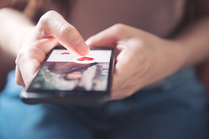 A close-up of a person's hand holding a smartphone, their thumb hovering over a screen showing a large heart icon, subtly hinting at online dating tips. In the background, the person's knees in jeans add a casual touch to this glimpse into digital romance.