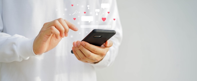 Person in a white shirt holding a smartphone with floating icons of hearts, chat bubbles, and search symbols above the screen, suggesting social media interaction and online dating tips. Blurred gray background.