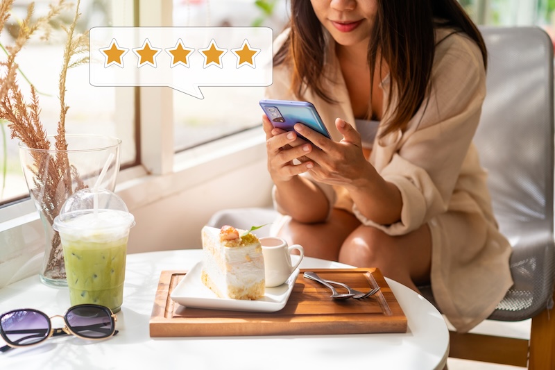 A woman in casual attire sits by a window, engrossed in her phone. On the table, a wooden tray holds cake and a small coffee cup alongside a green iced drink and sunglasses. With a five-star rating icon hovering above her, it's clear this spot excels in restaurant reputation management.