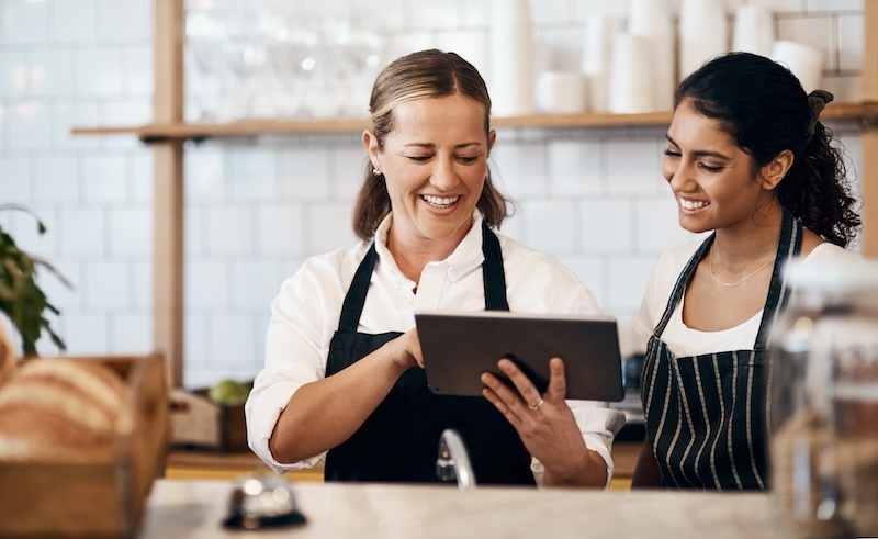 Two women in aprons stand in a bakery, smiling as they look at a tablet, perhaps discussing restaurant reputation management. The background features shelves with cups and a potted plant, while a basket of bread rests invitingly on the counter.