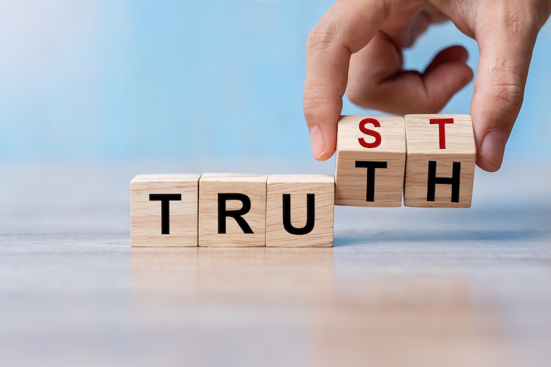 A person manipulates wooden blocks that spell "TRUTH," changing the "H" to an "S," transforming it into "TRUST." On a smooth surface, the scene subtly highlights how cultivating trust is essential to protect your brand reputation, illustrating the connection between truth and trust.