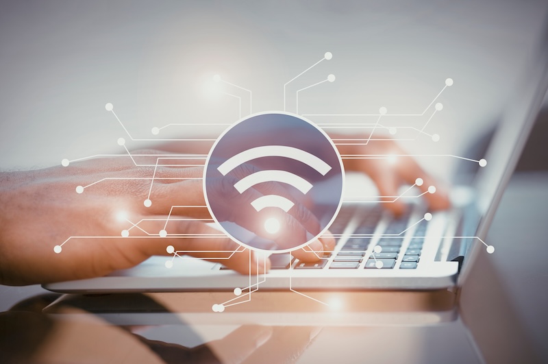 Close-up of a person's hands typing on a laptop keyboard. A large Wi-Fi symbol is superimposed over the image, surrounded by circuit-like lines, conveying the concept of network connectivity and how to protect your privacy online efficiently.