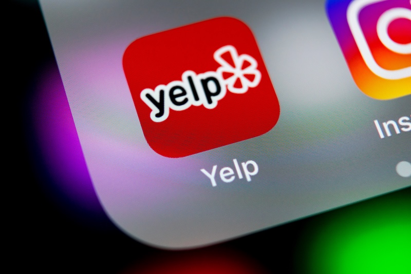 Close-up of a smartphone screen displaying the Yelp app icon. The red icon features "Yelp" and a stylized asterisk, with part of the Instagram app beside it. Dive into Yelp business reviews right from your phone to discover local favorites and hidden gems.