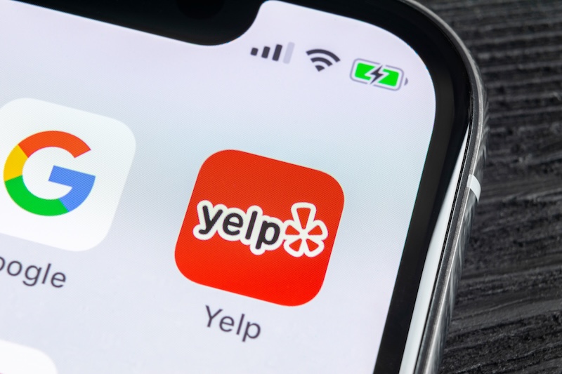 Close-up of a smartphone screen displaying app icons for Google and Yelp, where Yelp's icon with its red background and white asterisk symbolizes the gateway to countless business reviews. Battery and signal indicators are also visible at the top.