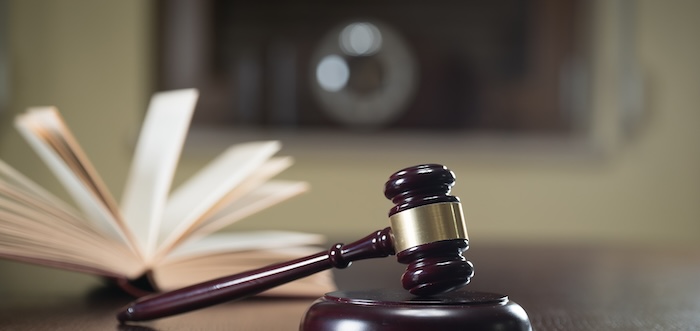 A wooden judge's gavel rests on a table beside an open book, with blurred background. The scene suggests a legal or judicial setting.