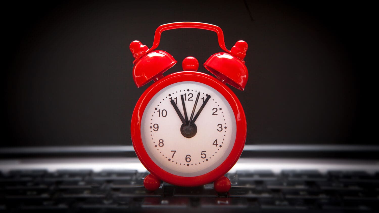 A red retro alarm clock sits on a computer keyboard. The clock shows the time as 12:00, with the hour and minute hands pointing straight up. The background is dark, emphasizing the bright red color of the alarm clock.