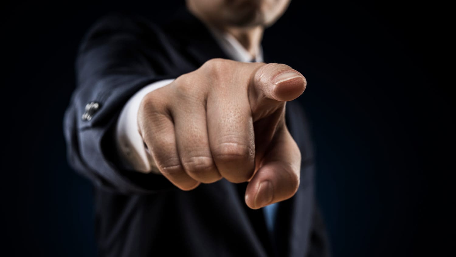 A person in a suit points a finger towards the camera against a dark background, creating a focused and dramatic effect.