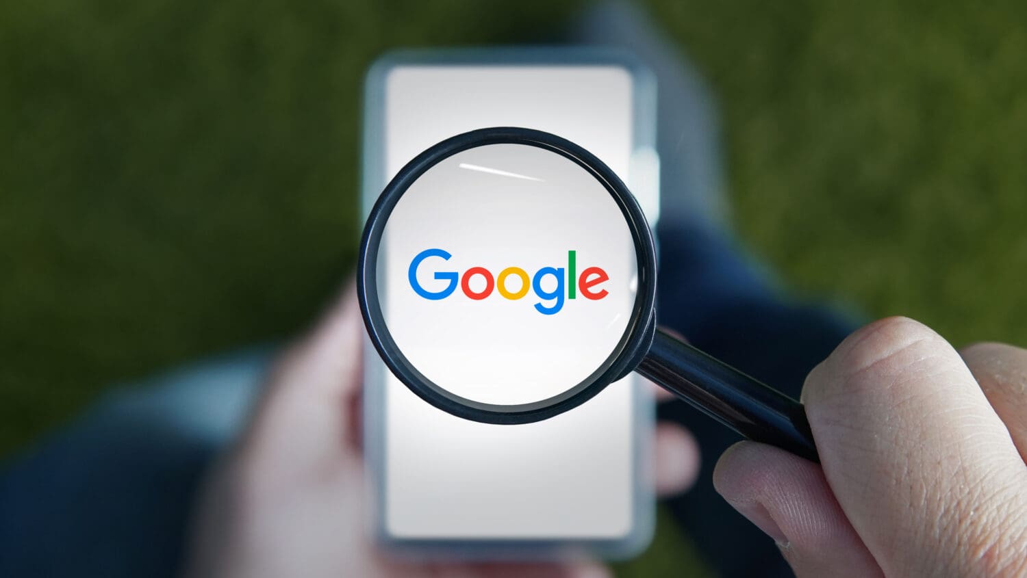 A person holds a magnifying glass over a smartphone displaying the Google logo. The background is blurred, and the logo appears magnified through the glass.