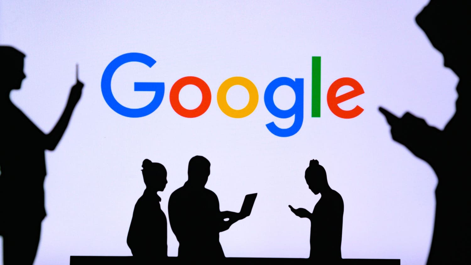 Silhouettes of four people using digital devices stand in front of a large screen displaying the colorful Google logo.