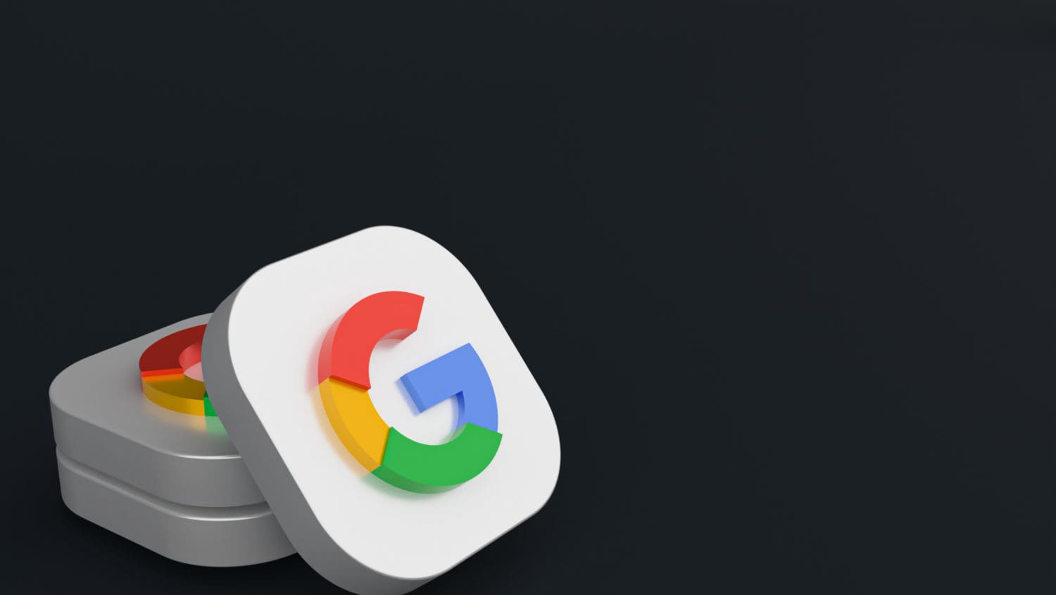 Two stacked, white, square objects each displaying a colorful "G" (Google logo) on their surfaces. The logos feature the colors blue, red, yellow, and green. The background is dark, creating a stark contrast with the bright logos.