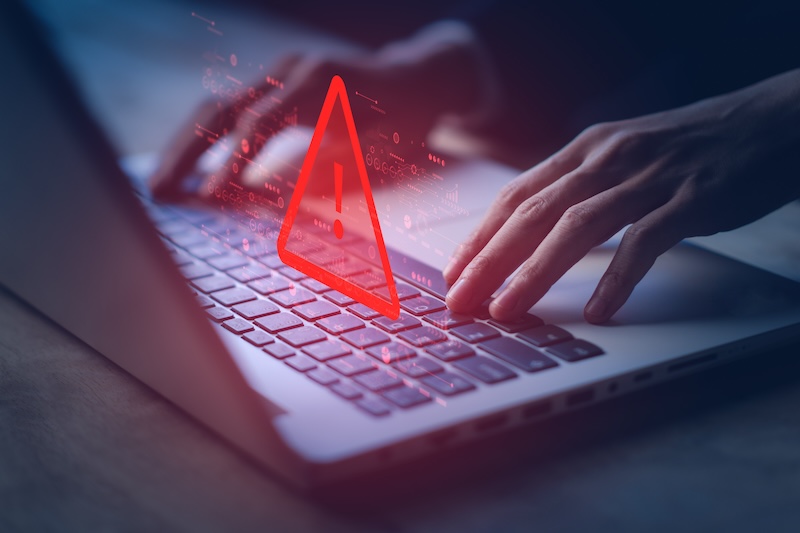 Hands typing on a laptop keyboard with a red warning triangle symbol containing an exclamation mark appearing on the screen, indicating a security alert or error message related to IDTrue.