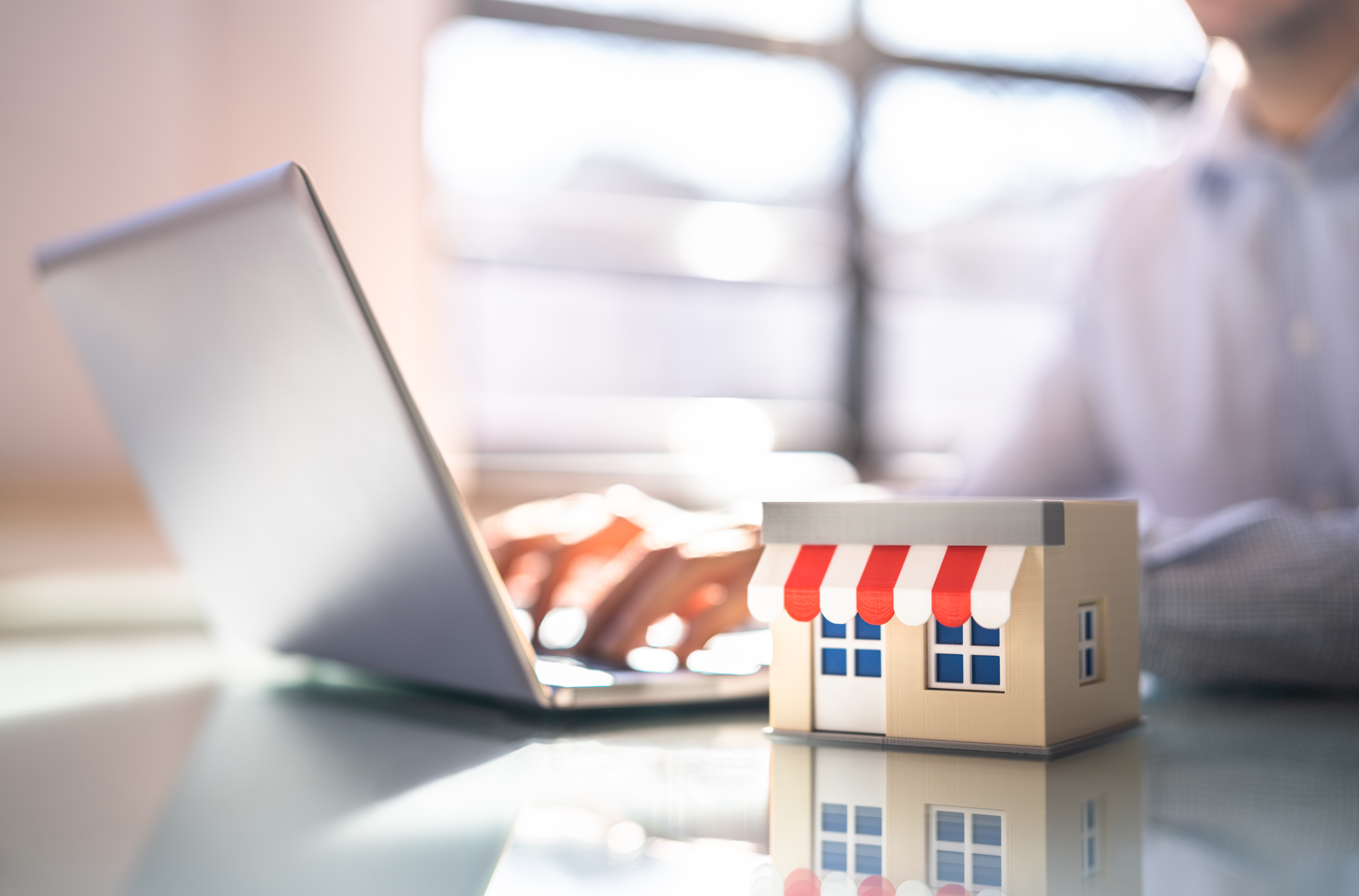 A person uses a laptop on a clear desk, focusing on their work. In the foreground, there is a small model of a storefront with a red and white awning. Sunlight streams through the window in the background.