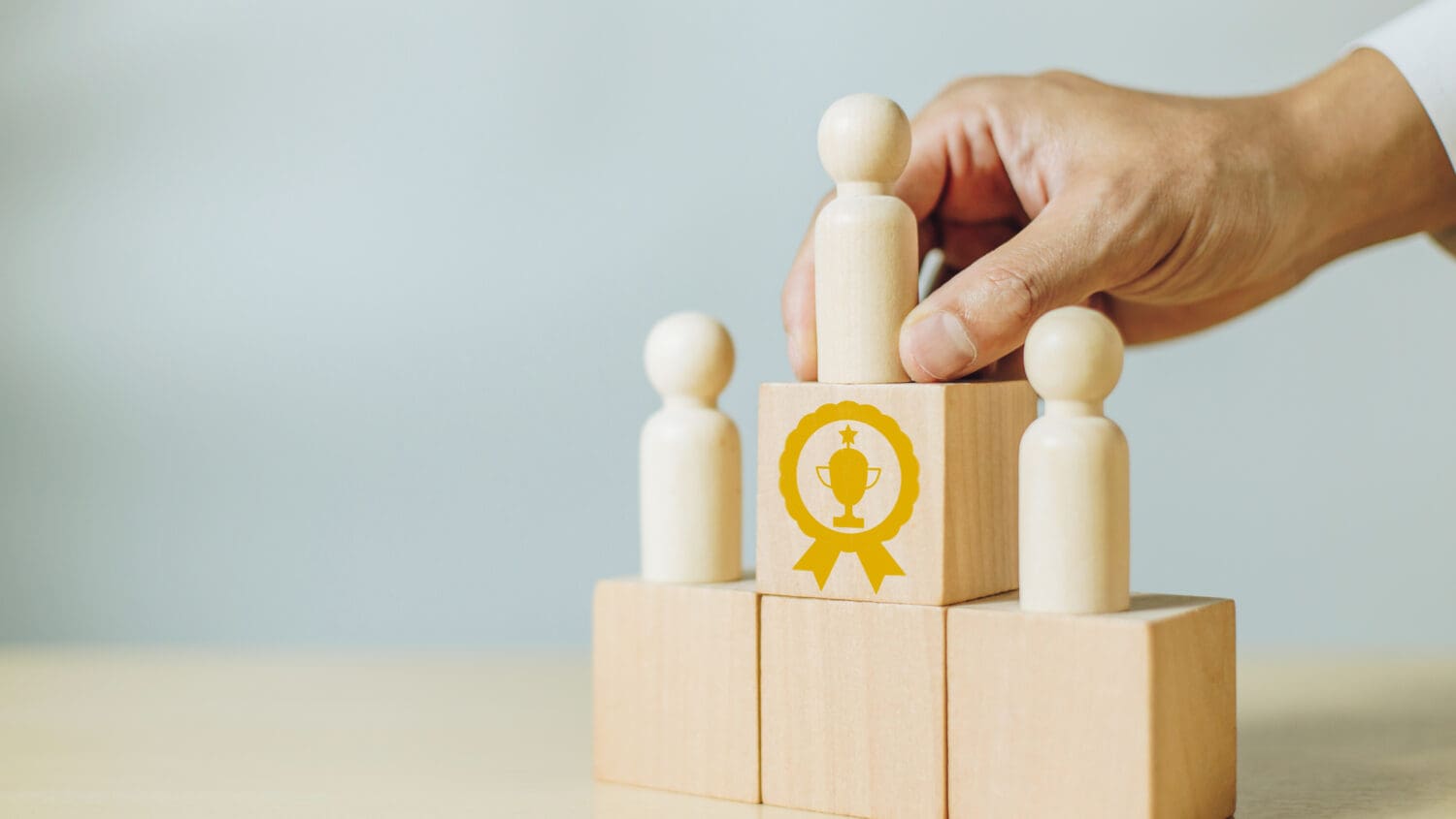 A hand places a wooden figure on a podium made of wooden blocks. One block has a yellow trophy symbol, indicating first place. Two more figures stand on lower blocks, representing second and third places.