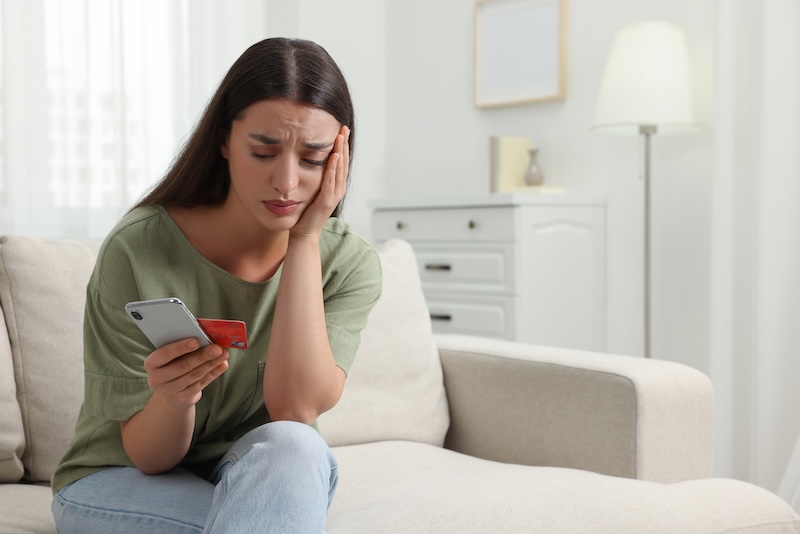 A woman with long, dark hair sits on a couch, looking distressed while holding her hand to her head and staring at her phone and a credit card. She wears a green shirt and jeans. The background features light-colored furniture and a lit floor lamp as she contemplates whether to opt out of an offer.