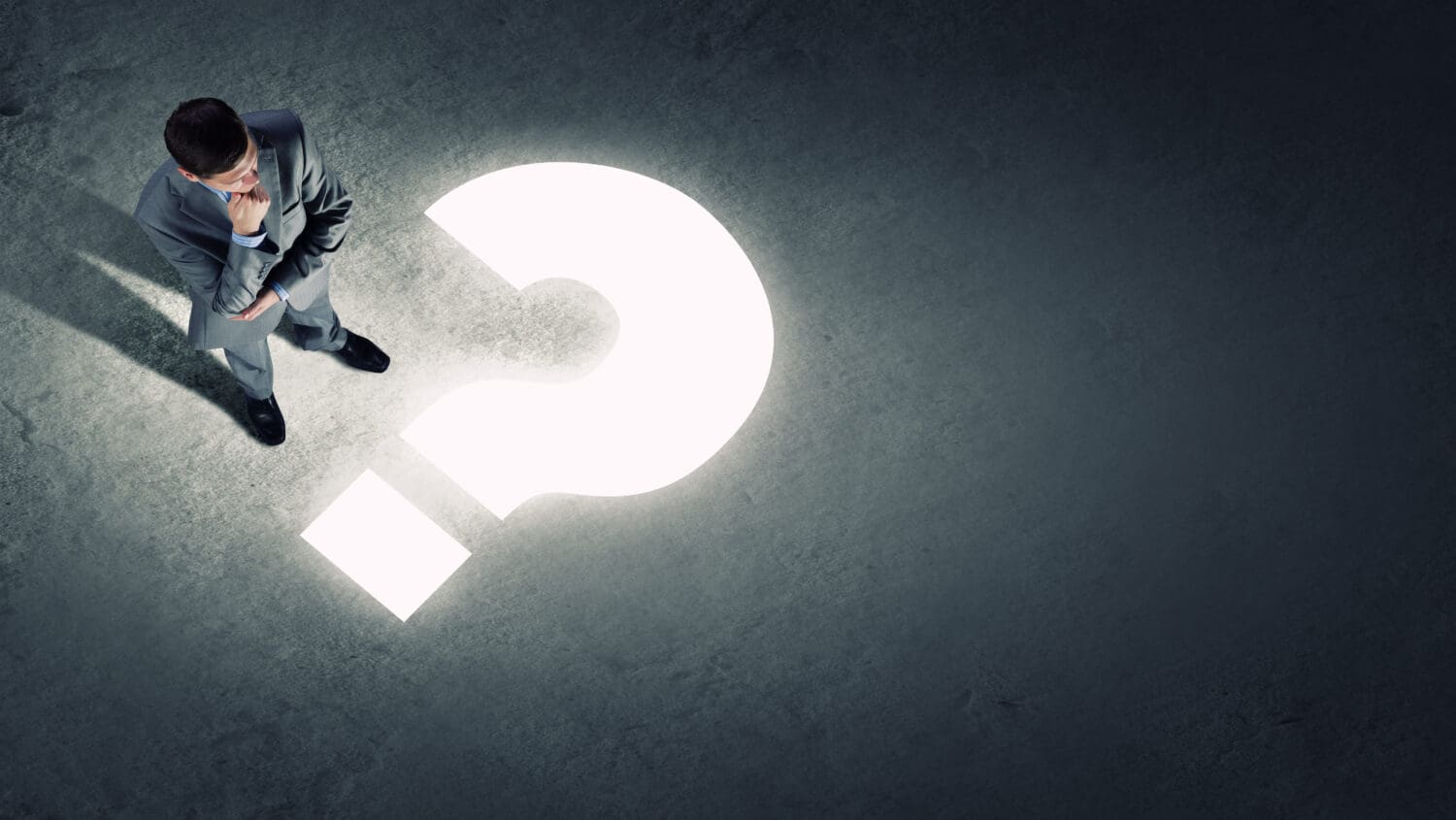 A person in a suit stands on a dark surface, looking down thoughtfully at a large, glowing white question mark projected on the ground. The space around them is shadowy and undefined.