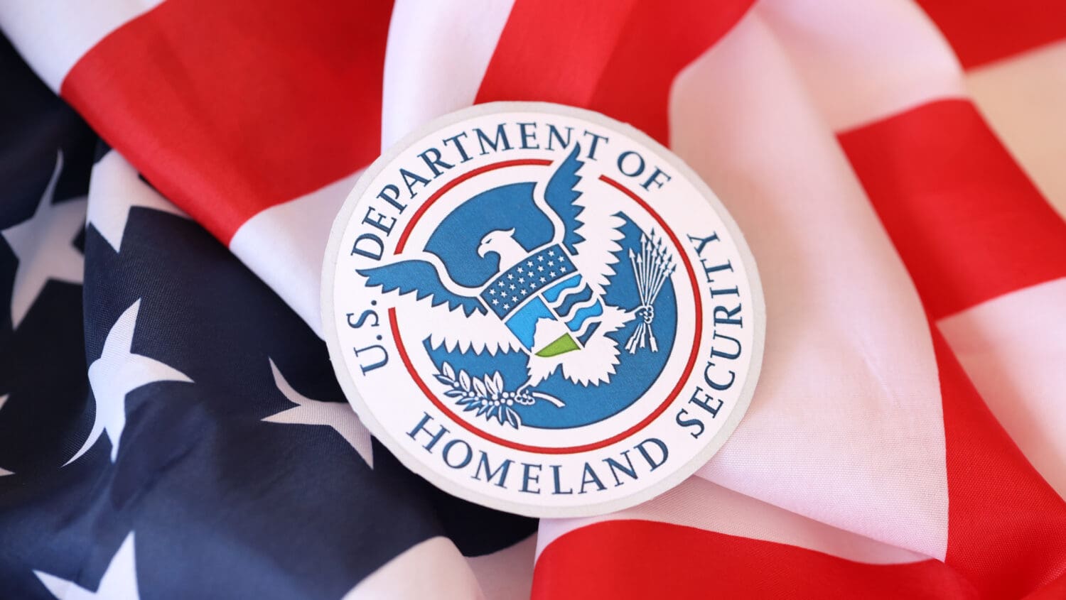 Seal of the U.S. Department of Homeland Security on an American flag. The seal includes an eagle, shield, and olive branches, symbolizing protection and peace.