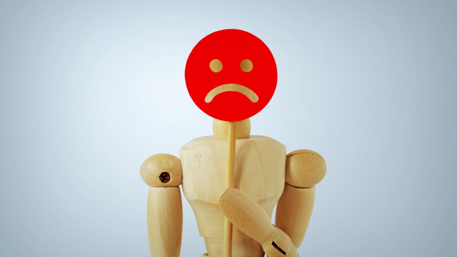 A wooden mannequin holds a stick with a red circle featuring a white sad face, symbolizing unhappiness. The background is pale blue, giving the scene a simple and neutral look.