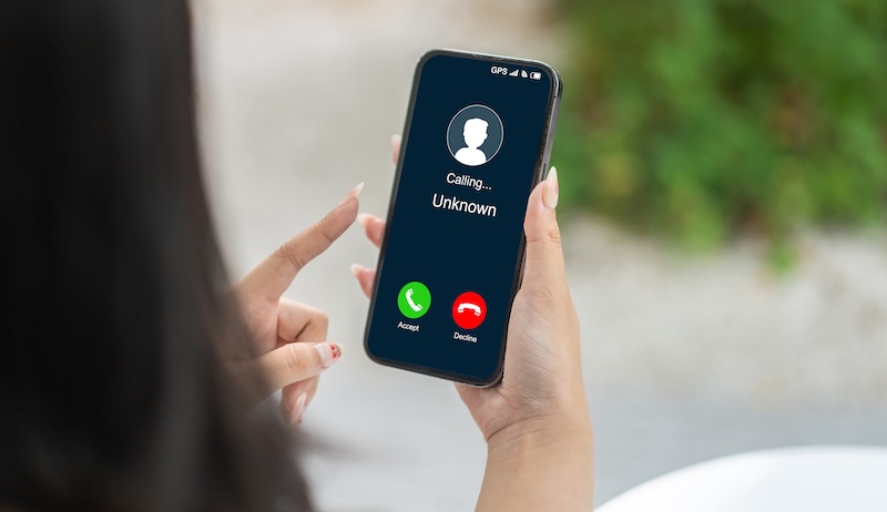 A person holds a smartphone displaying an incoming call from an unknown number. The screen shows "Calling... Unknown" with green "Accept" and red "Decline" buttons, hinting they may want to opt out of this mystery. The background is blurred with hints of greenery.