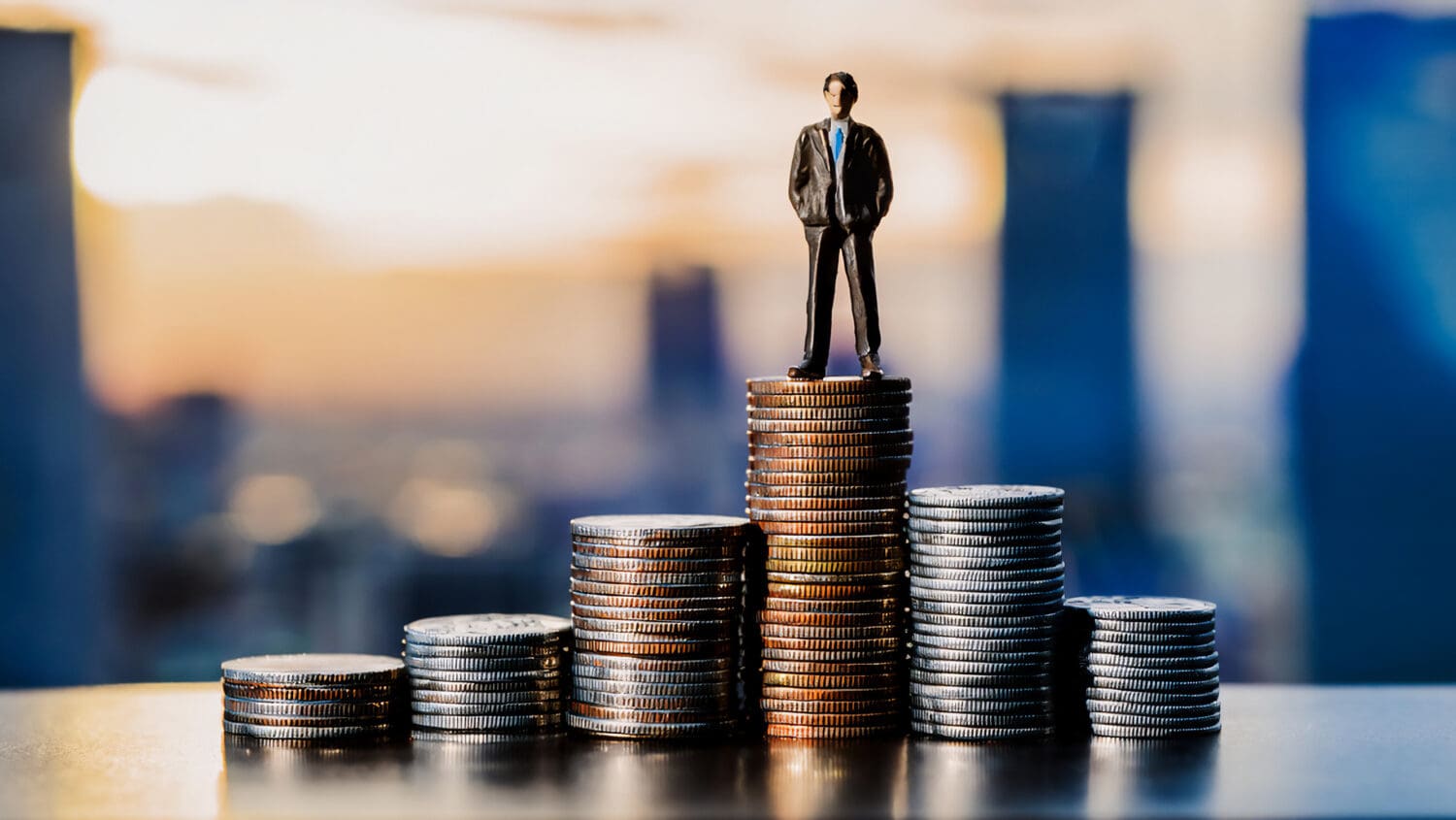 A miniature figure of a man in a suit stands on top of a stack of coins, with smaller stacks decreasing in height beside him. The background is blurred, suggesting an urban landscape at sunset.