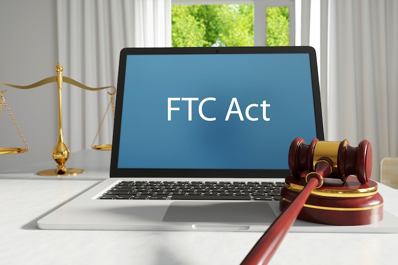 A laptop displays "FTC Act" on the screen, situated on a desk with a wooden gavel and a set of brass scales in the background, suggesting a legal or regulatory theme related to the Federal Trade Commission.