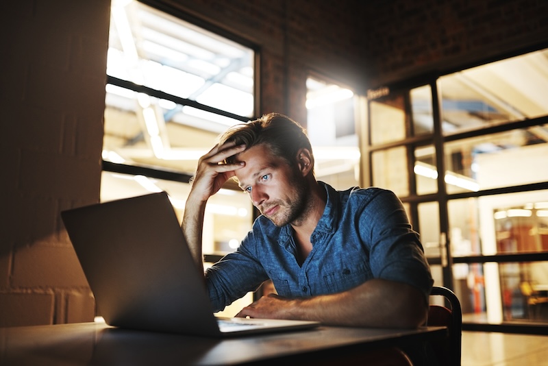 A man in a denim shirt looks frustrated while sitting at a desk with a laptop. He rests his head on his hand, staring at the screen. The room has industrial-style glass doors and ceiling lights.