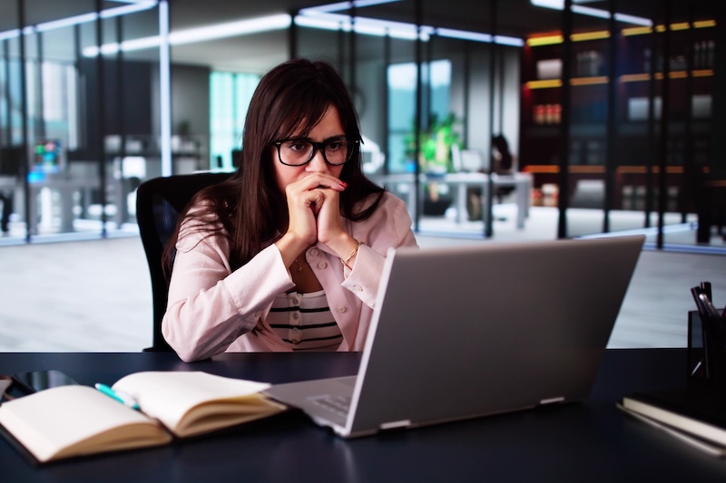 A woman with long dark hair and glasses sits at a desk, looking intently at a laptop screen. Open notebooks are on the desk in front of her. The background shows a modern office with glass walls and shelves, as she delves into a report on online extortion.