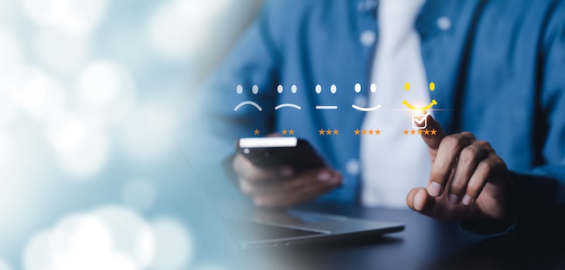 A person in a blue shirt engages with a digital satisfaction scale on a touch screen, an essential tool for online presence management. Emojis range from sad to happy, and the individual selects the happiest emoji, marked by a yellow checkmark and five stars.