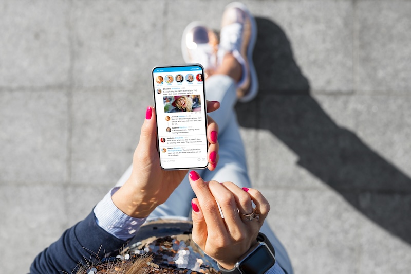 A person with pink nail polish casually scrolls through their social media feed, the smartphone's glow highlighting a series of images and text posts. Standing on a concrete surface, their shadow stretches out, unaware of potential online extortion lurking in the digital shadows.