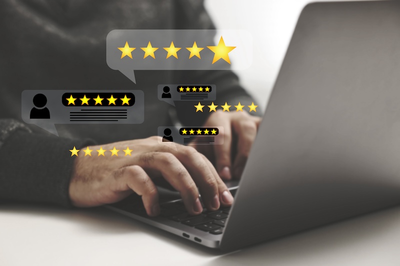 A person typing on a laptop with floating customer review icons, including multiple five-star ratings and comment bubbles. The scene suggests online feedback or evaluation.