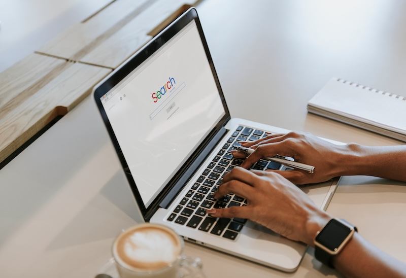 Person typing on a laptop displaying a search engine on the screen. A notebook lies beside the laptop on the white desk. A cup of coffee with a frothy top sits in the foreground.