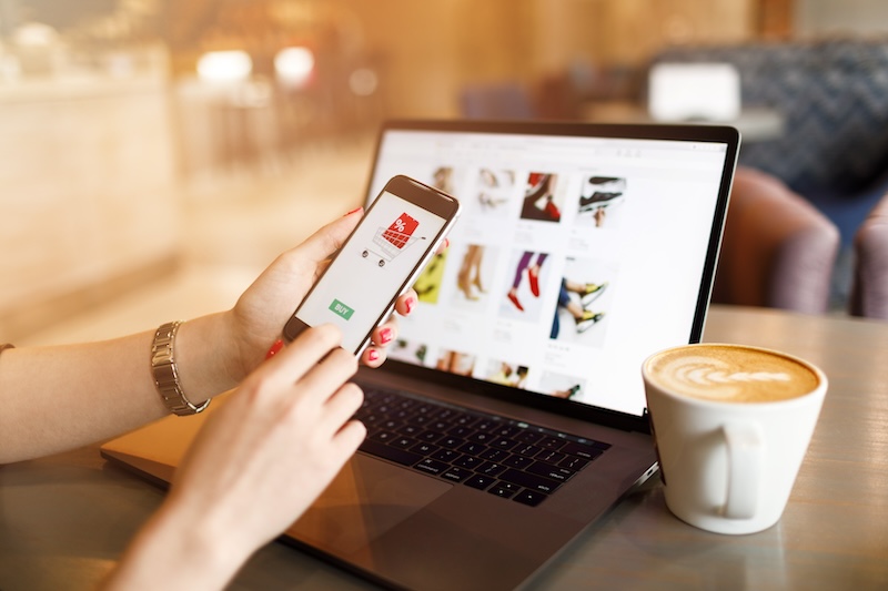 A person holds a smartphone displaying a shopping app while sitting at a table with a laptop. The laptop screen shows an online store with images of shoes. A cappuccino in a mug is next to the laptop. The setting is cozy and well-lit.