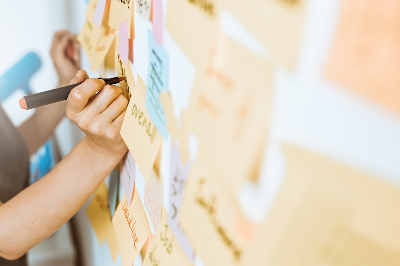 A person writes on sticky notes attached to a wall. The notes are arranged in a collage of various colors, including pink, yellow, and blue. The scene suggests brainstorming or planning activity.