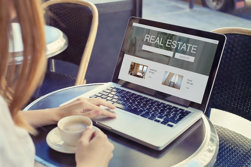 A person at an outdoor cafe table holds a cup of coffee while using a laptop. The screen displays a real estate search page with property listings. Nearby, empty chairs and a sunlit atmosphere suggest a casual setting.