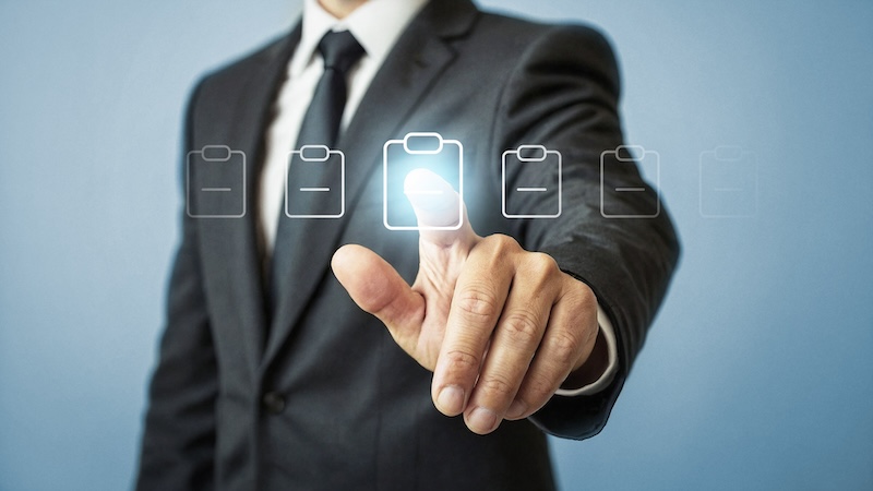 A man in a suit and tie standing against a blue background is selecting a glowing digital icon of a clipboard with his finger, while other similar, non-glowing clipboard icons are lined up beside it.