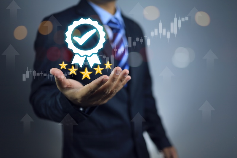 A person in a suit and tie holds out their hand with a holographic image of a quality certification badge displaying a checkmark and five stars, symbolizing the importance to brandyourself. The background features abstract financial graphs and arrows indicating a positive trend.