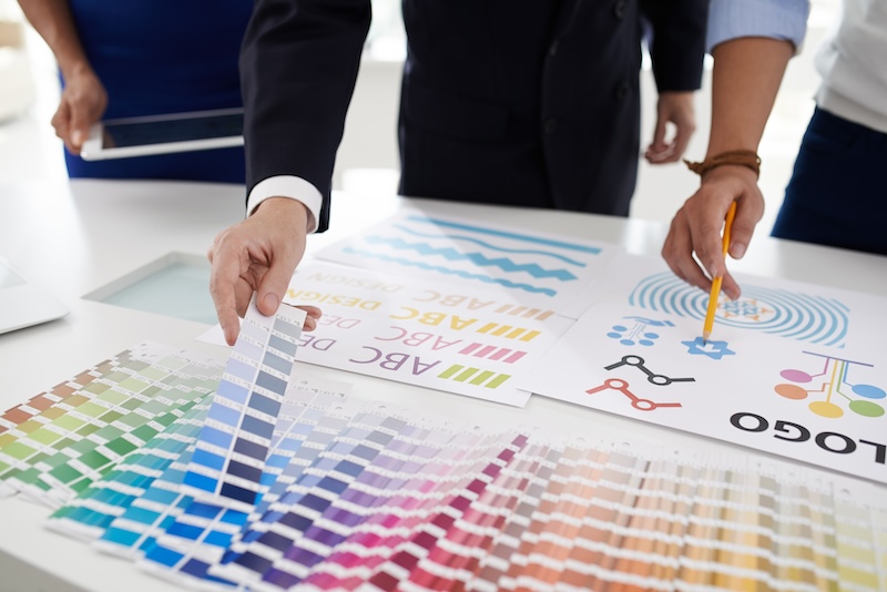 Close-up of people at a table reviewing color swatches and design materials, crucial for corporate reputation management. Two hands are visible pointing at various design samples, one holding a pencil and the other touching a palette of colors. Various printed graphics and charts are spread across the table.