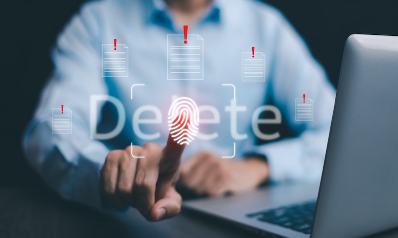 A person touching a digital fingerprint icon on a transparent screen with the word "Delete" in the background. Red exclamation marks appear on document icons above the fingerprint, suggesting a digital deletion or security action. A laptop is visible on the right.