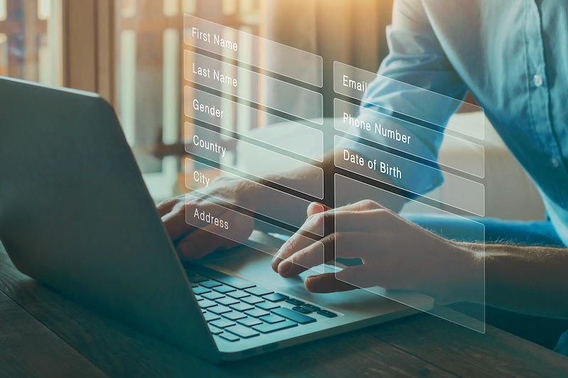 Person typing on a laptop with transparent graphical overlays displaying form fields such as First Name, Last Name, Gender, Country, City, Address, Email, Phone Number, and Date of Birth. Sunlight streams through a window in the background.