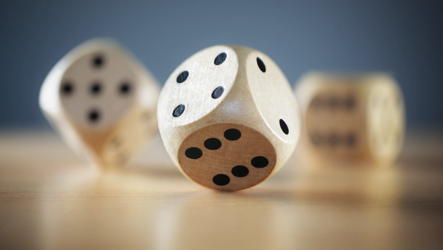 Three wooden dice, reminiscent of fortune's most admired companies, are placed on a smooth wooden surface. One die is in focus, displaying a five, while the others are slightly blurred in the background against a muted blue backdrop, resting on different faces.